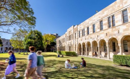 people walk and sit on grass by stone buildings with an arched walkway